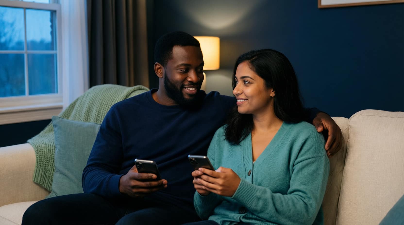 A couple sitting close together on a sofa at home, each holding their own phone mid-conversation, looking thoughtful and at ease.